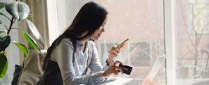 A woman sits in front of a window looking at her cell phone and holding her credit card.