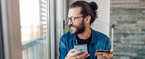 A smiling man looks out the window while holding his smartphone and credit card.