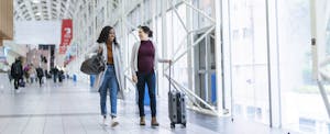 Two friends on their way to Canada walk side by side in an airport with their luggage.