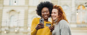 Two smiling women stand side by side looking at a smartphone, while one holds her United Quest Card in her other hand.