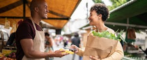 A woman holding a bag of produce and a vendor smile at each other at an outdoor market as she uses her mobile credit card to pay.