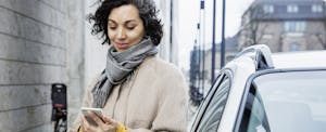 A woman standing next to her car smiles while looking at her phone because she received her car insurance refund.