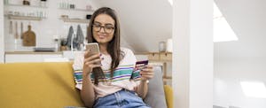 Young woman on her sofa, shopping online with her mobile phone, paying with her Credit One Bank Platinum Visa for Rebuilding Credit