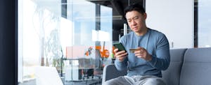 A man with a concerned expression holds his debit card and uses his smartphone to learn about how to protect yourself from debit card fraud.