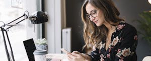 A woman seated at a desk uses her smartphone to research wire transfer vs direct deposit to understand the differences between the two.