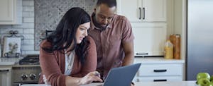 A couple stand at their kitchen counter using a laptop to shop for new car insurance after some insurers drop hyundai and kia models.