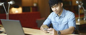 A person seated at a desk in front of a laptop holds their debit card while using their smartphone.