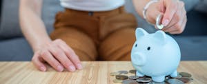 A little blue piggy bank on a table is sitting in the middle of a pile of coins, while a hand drops a coin in its slot.