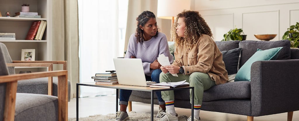 Two women sit on the couch with an open laptop, discussing finances.