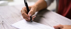 Close-up photo of a person's hands as they write a check on a wooden tabletop.