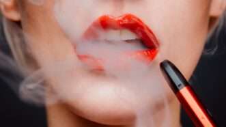 Woman with red lipstick has vapor coming out of her open mouth, with a vape pen near her lips. | Photo by <a href="https://unsplash.com/es/@chiarasummer?utm_source=unsplash&utm_medium=referral&utm_content=creditCopyText">Chiara Summer</a> on <a href="https://unsplash.com/s/photos/vaping?utm_source=unsplash&utm_medium=referral&utm_content=creditCopyText">Unsplash</a>