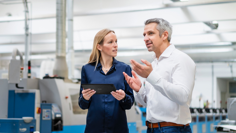 Two manufacturing employees are duscussing together in a facrory and one of the employee is holding an ASUS ExpertBook B3 detachable laptop.