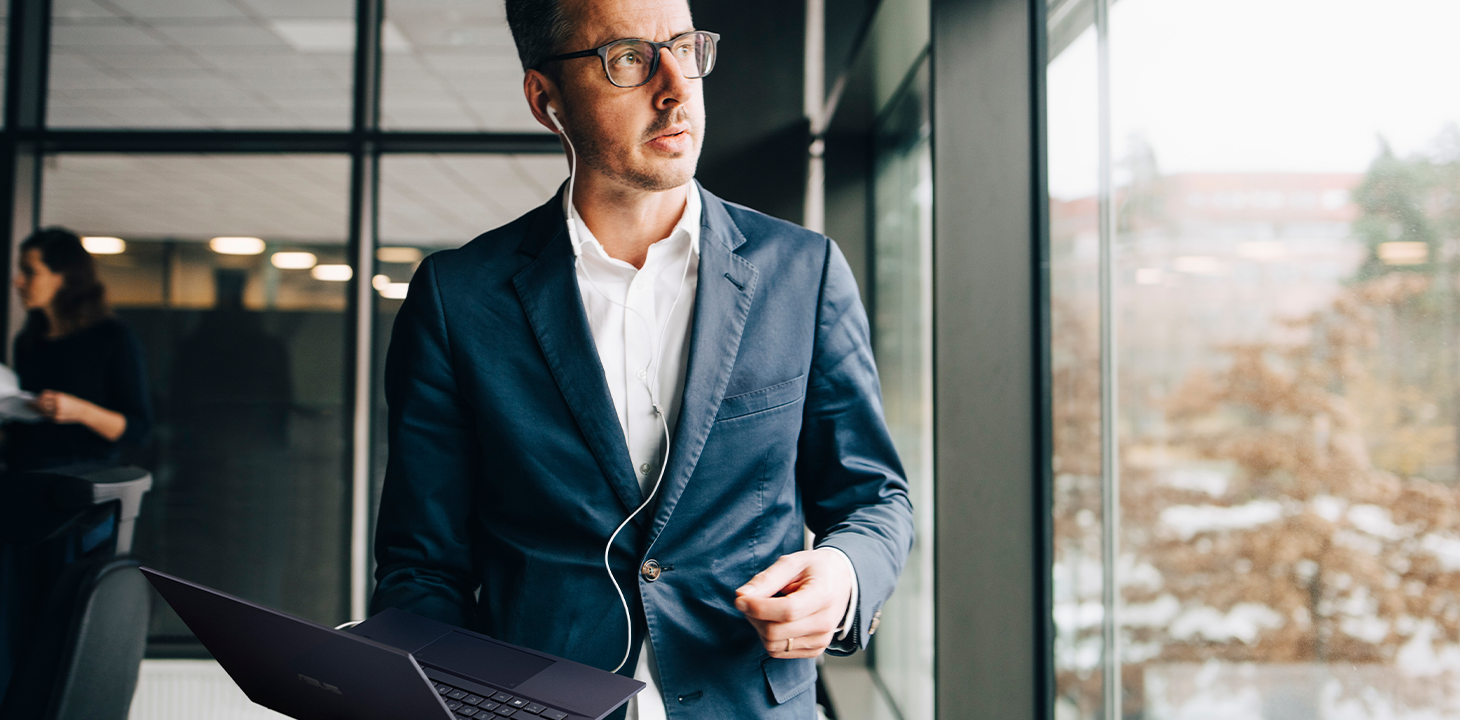 An executive is in a video-conferencing, holding ASUS ExpertBook laptop on hand, standing by the window in office.