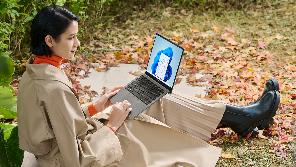 A woman sitting on the ground in a forest holding an ASUS Zenbook S 13 OLED laptop on her laps