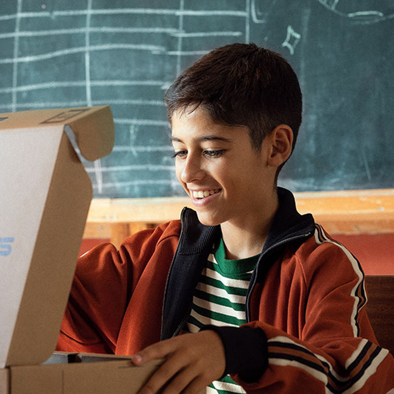 A smiling student opening an ASUS laptop box while sitting in front of a blackboard in a classroom