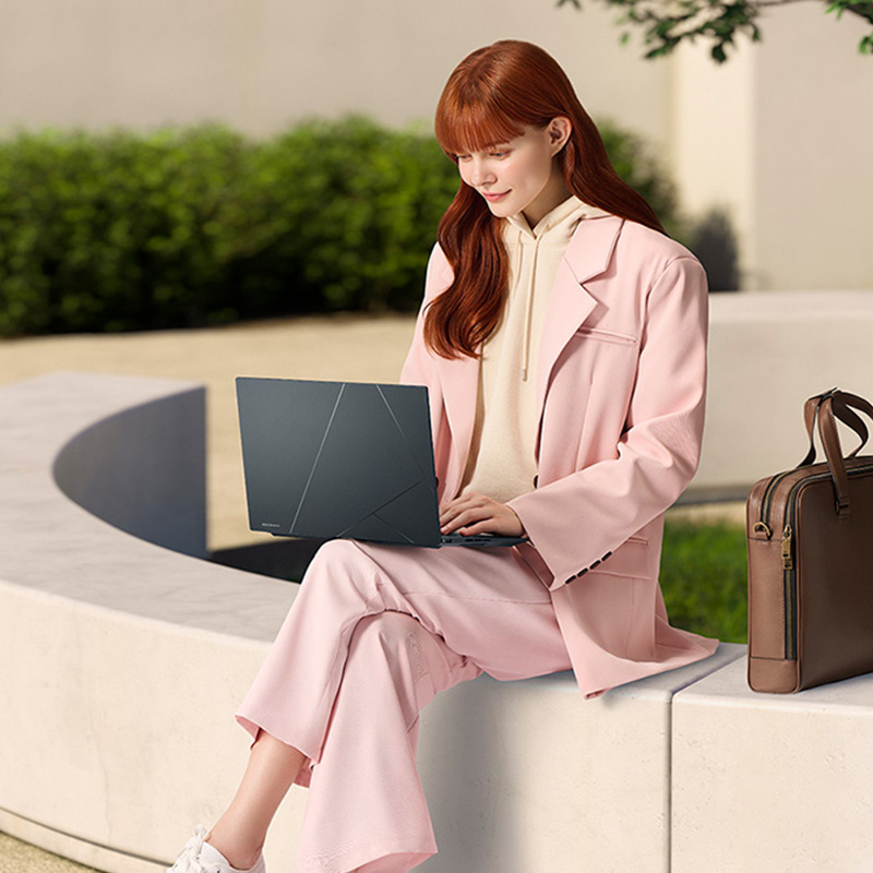 A woman sitting on a wall with an ASUS Zenbook 14X OLED laptop on her laps and a brown briefcase next to her with a tree in the background