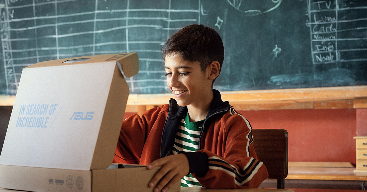 A smiling student opening an ASUS laptop box while sitting in front of a blackboard in a classroom