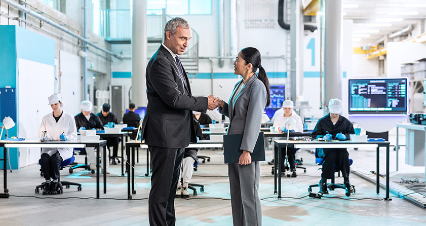 A man in a suit shaking hands with a woman wearing a suit, holing an ASUS laptop while standing in front of working technical staff