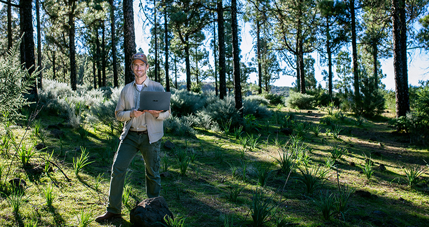 a young man smiling while standing in a sunlit forest and holing an ASUS laptop?