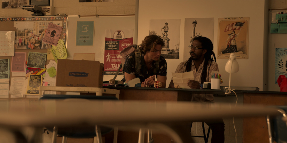 John B stands next to Mr. Sunn at his desk in the classroom.