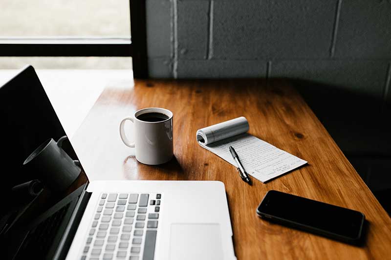 A laptop, coffee, notebook and cup of coffee on the surface of a desk.