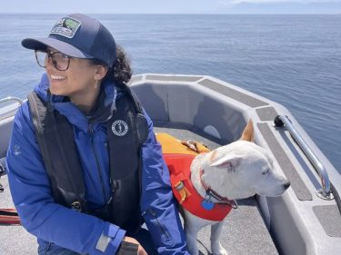 Aisha Rashid, seated left and Eba a white dog in a life jacket, standing right, on a small boat in Puget Sound on a sunny day.