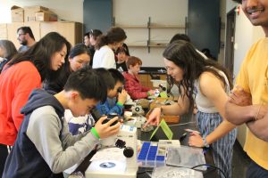 Kids look at different types of rocks through a microscope, assisted by graduate student Haskelle White-Gianella.