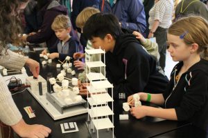 A young kid builds a structure with marshmallows and sticks. The structure sits on a plate that moves, simulating an earthquake, to see if the structure is strong enough to stay together.