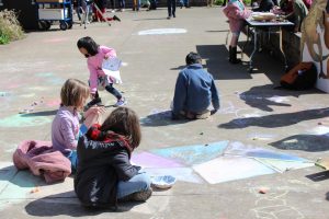 Four young kids sit near each other on the concrete working on chalk designs.