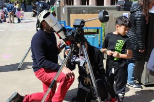 An adult kneels to look into a telescope with Professor Baptiste Journaux to see the sun's surface.