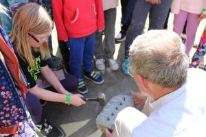 A young kid wearing safety glasses holds a small sledgehammer to hammer a row of pegs into a large rock to split it down the middle.