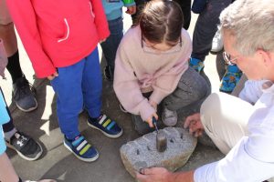 A young kid wearing safety glasses holds a small sledgehammer to hammer a row of pegs into a large rock to split it down the middle.