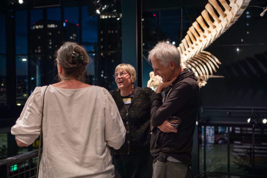 Department community members socializing at the ESS 130th Anniversary Celebration in the Burke Museum in front of a dinosaur fossil.