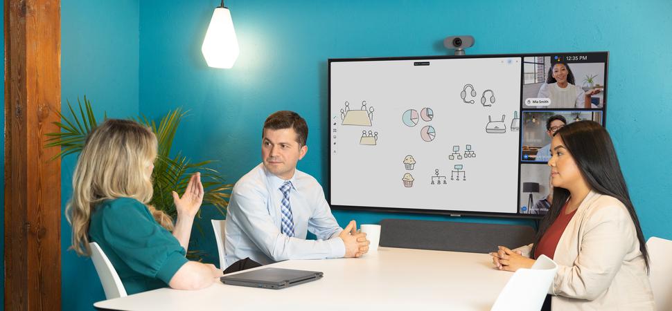 An image of a group of people in a meeting room with a Google Meet Series One Small Room mounted to the wall at the head of the table.