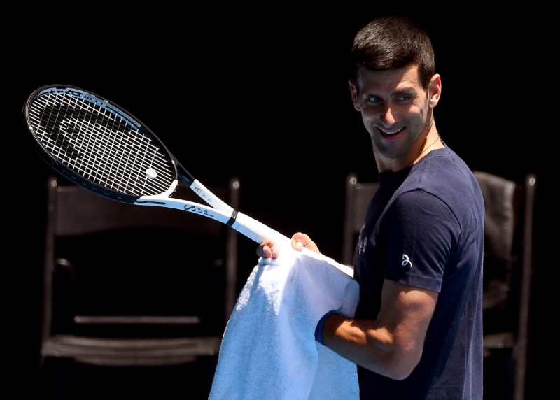 Djokovic smiles while holding a tennis racket and towel.