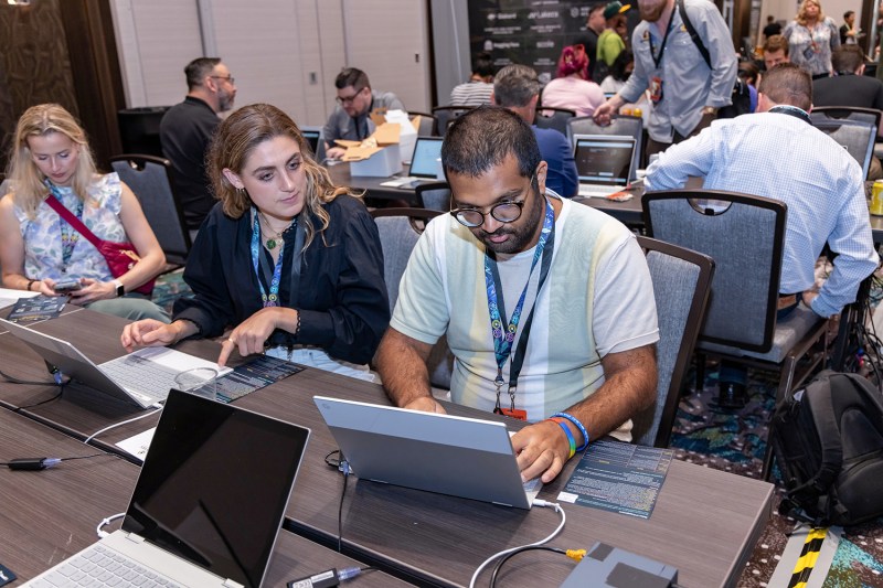 Two adults wearing matching lanyards designating them as volunteers sit next to each other at a table with laptops in front of them. One of the volunteers leans to the side to look at the other's screen as he types. Behind the volunteers, other people sit at tables with laptops and phones.