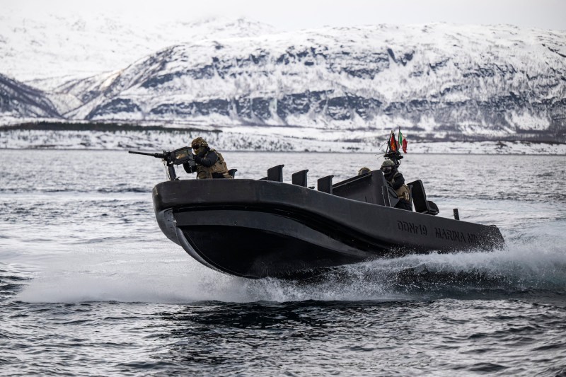 Three members of the Italian Marines in helmets and bullet-proof vests stand onboard a small motorized military boat as it cuts through the water in front of a snow-covered ridge of land. The soldier at the bow aims a rifle, while the other two sit at the stern and steer the vessel.