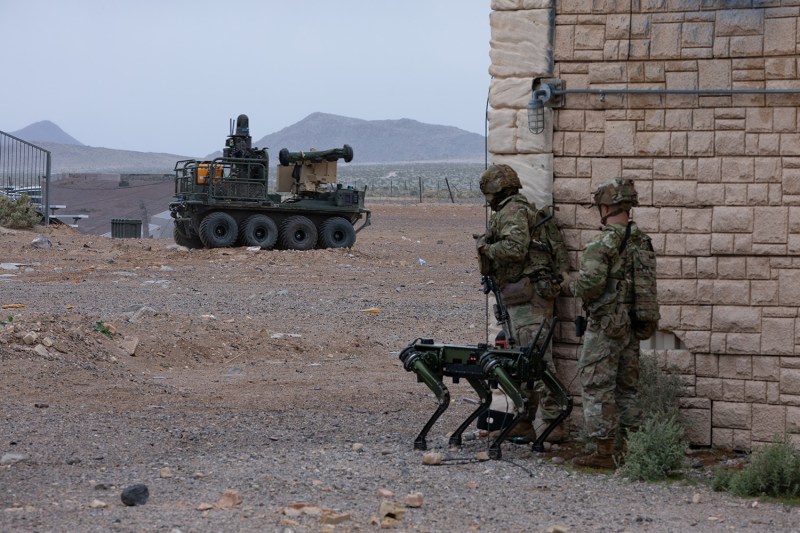 Two soldiers in combat gear carefully peer around the corner of a wall while a robotic dog stands at their knees. They look across a desert landscape toward a robotic vehicle in the distance.