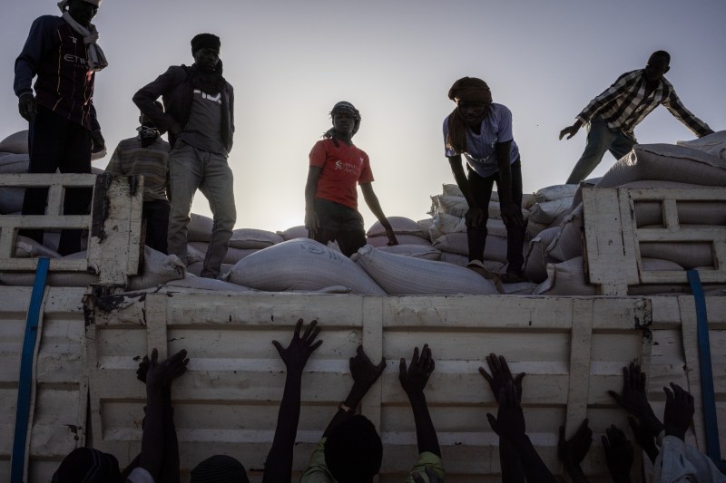 Arms reach up to sacks of aid as they are unloaded by people atop them on a truck above.