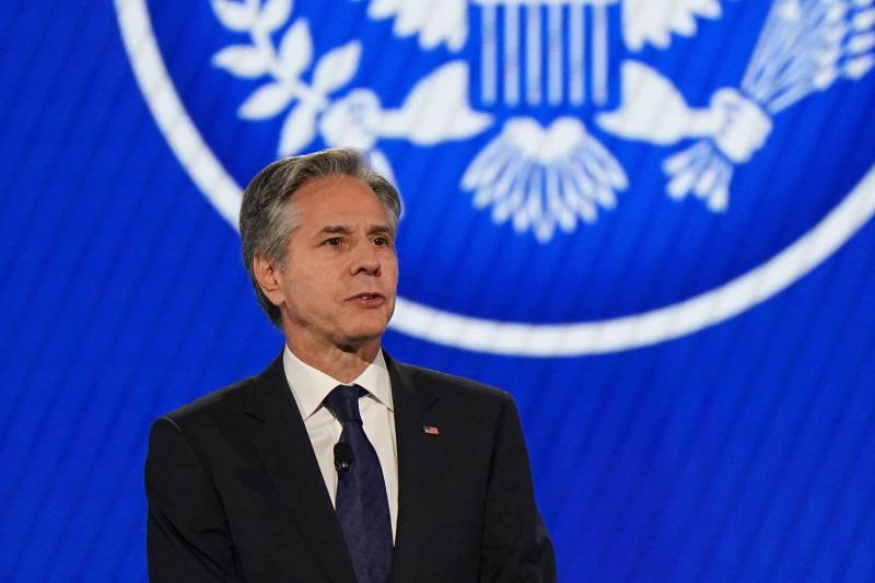 U.S. Secretary of State Antony Blinken, a man in his 60s dressed in a black suit and navy blue tie, stands in front of a blue digital background featuring a U.S. government seal logo. as he speaks onstage during a conference.