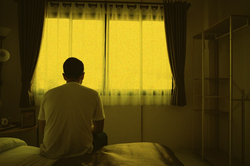 A man in a white T-shirt sits on the edge of a bed looking out a window, the view outside partly obscured by hazy curtains. A haze of yellow light covers entire scene.