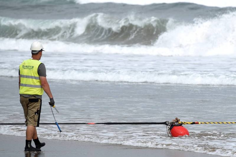 A person in a construction vest and hard hat walks toward the shoreline where a thick cable lies.