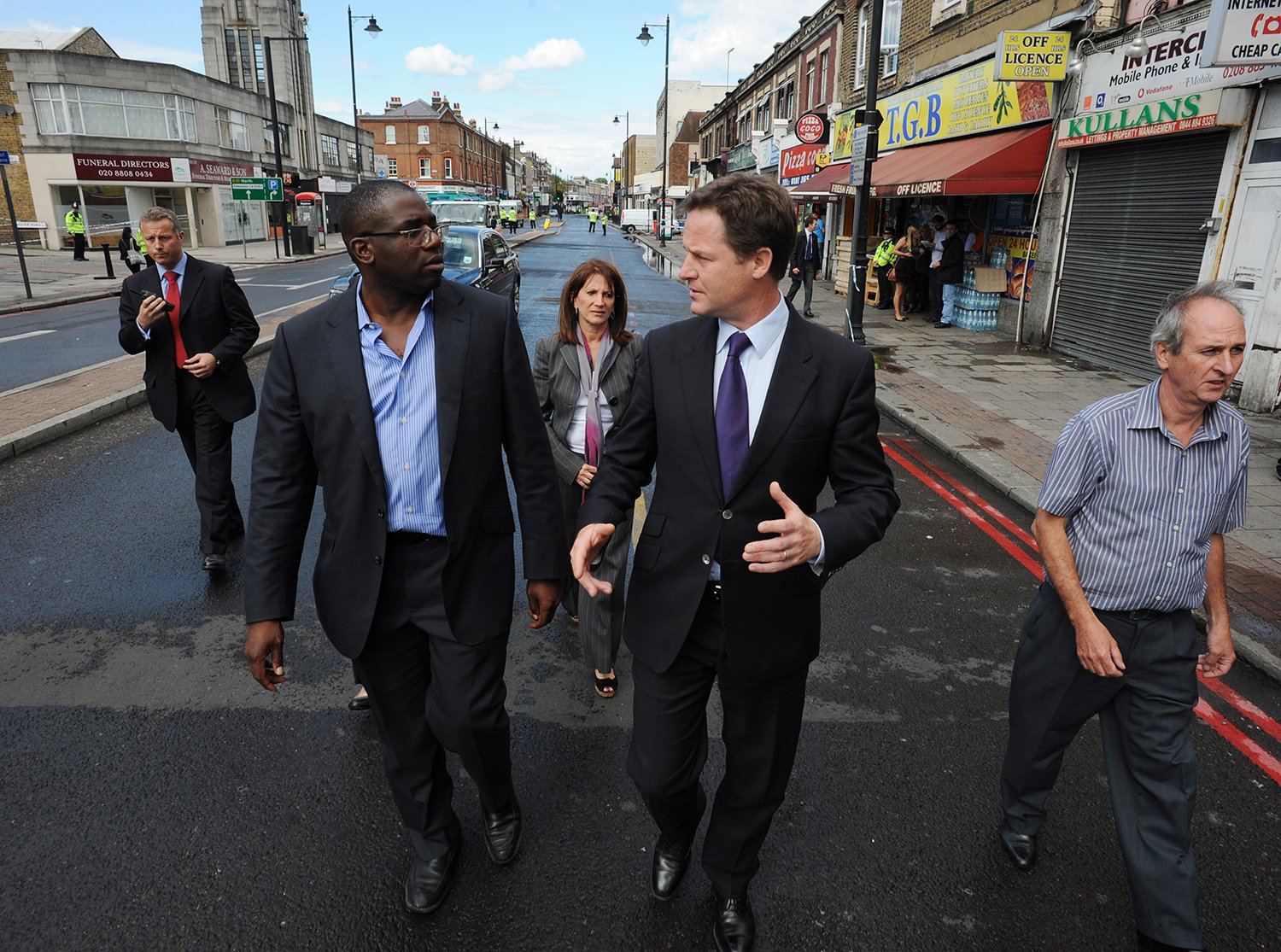 David Lammy wearing a suit with no tie and Nick Clegg, gesturing in a suit and tie, walk with local residents down a Tottenham street lined with shops.