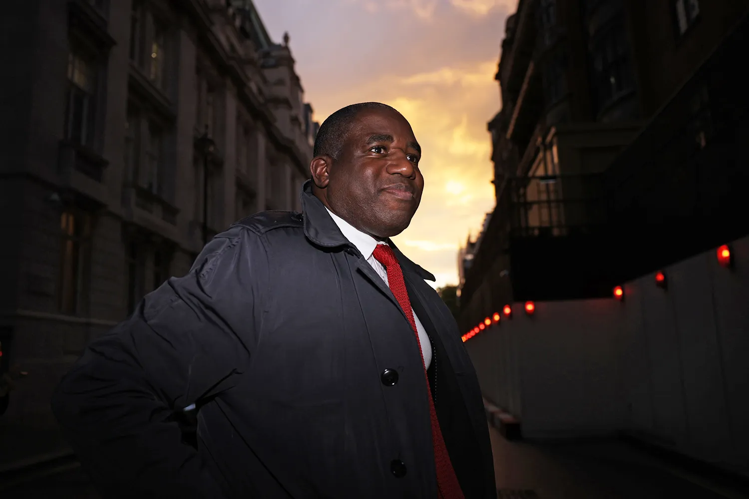 A man wearing a dark overcoat and red tie smiles as he crosses a street between buildings against a glowing sky.