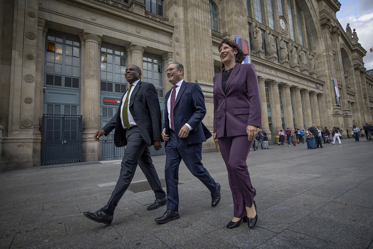 Two men and a woman, all in suits, walk down a Paris street past a train station where people with suitcases exit the building in the background.