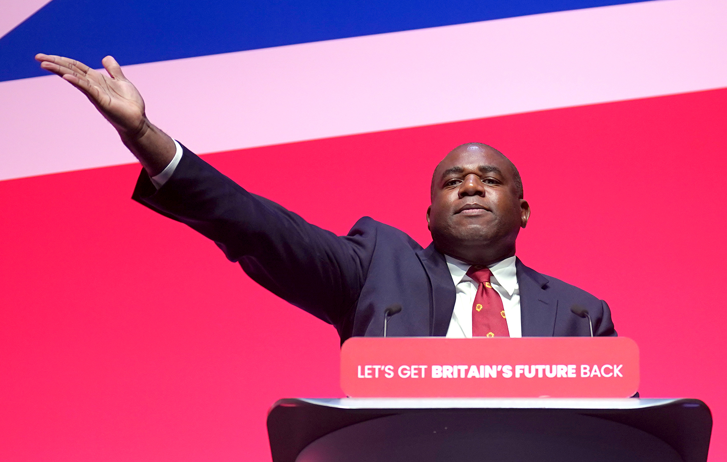 David Lammy wearing a dark suit and tie gestures with one hand as he stands behind a lectern that reads: "Let's Get Britain's Future Back."