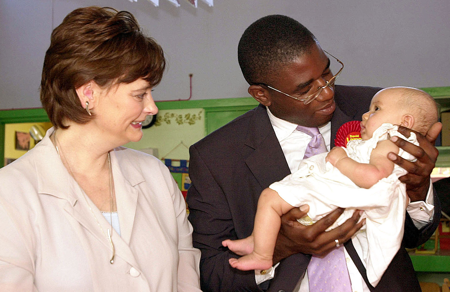 A woman in a beige jacket smiles as she looks at a man in glasses and suit and tie holding a small baby in a white dress.