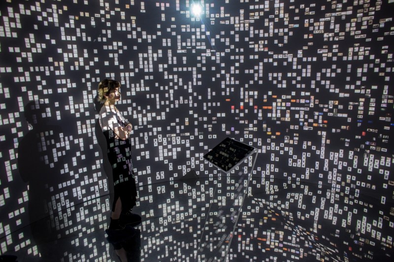 A woman stands in the middle of a room with black walls with tiny images of historical photographs and documents projected onto every corner of the room.