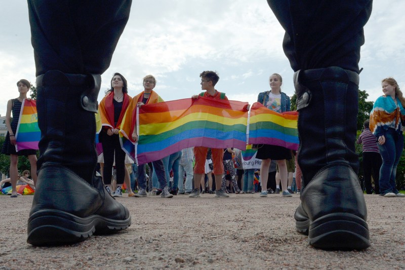 A view from the ground level shows people across a clearing hold rainbow flags during a gay pride rally. The viewer is poised just behind the back of a person standing in front of the demonstrators, viewing them from between that person's two black boots.