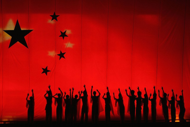 Silhouettes of undergraduates dance on a stage with a giant Chinese flag in the background.